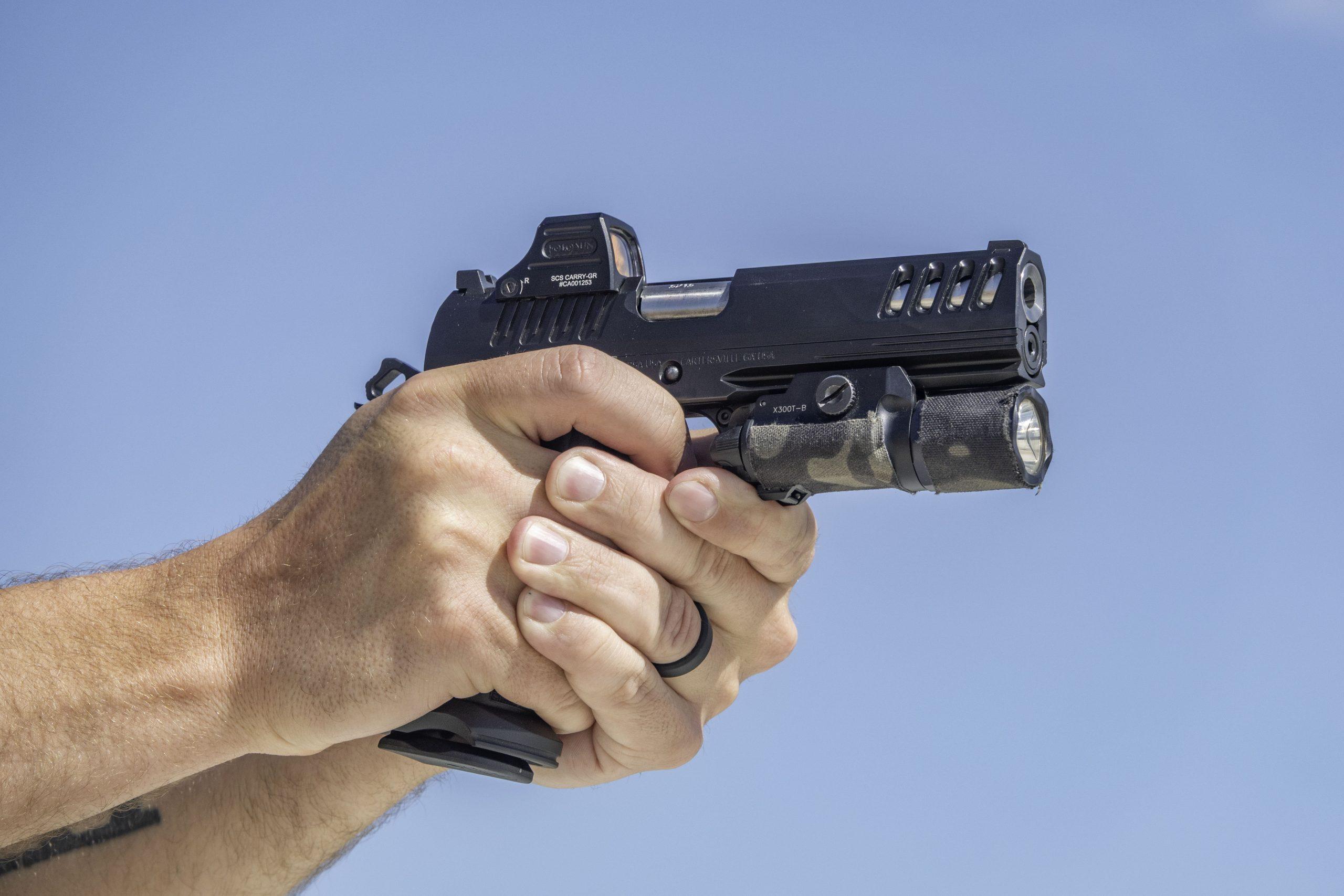 Close-up of two hands gripping the Bersa MX2I black pistol with a weapon light, set against a clear blue sky.